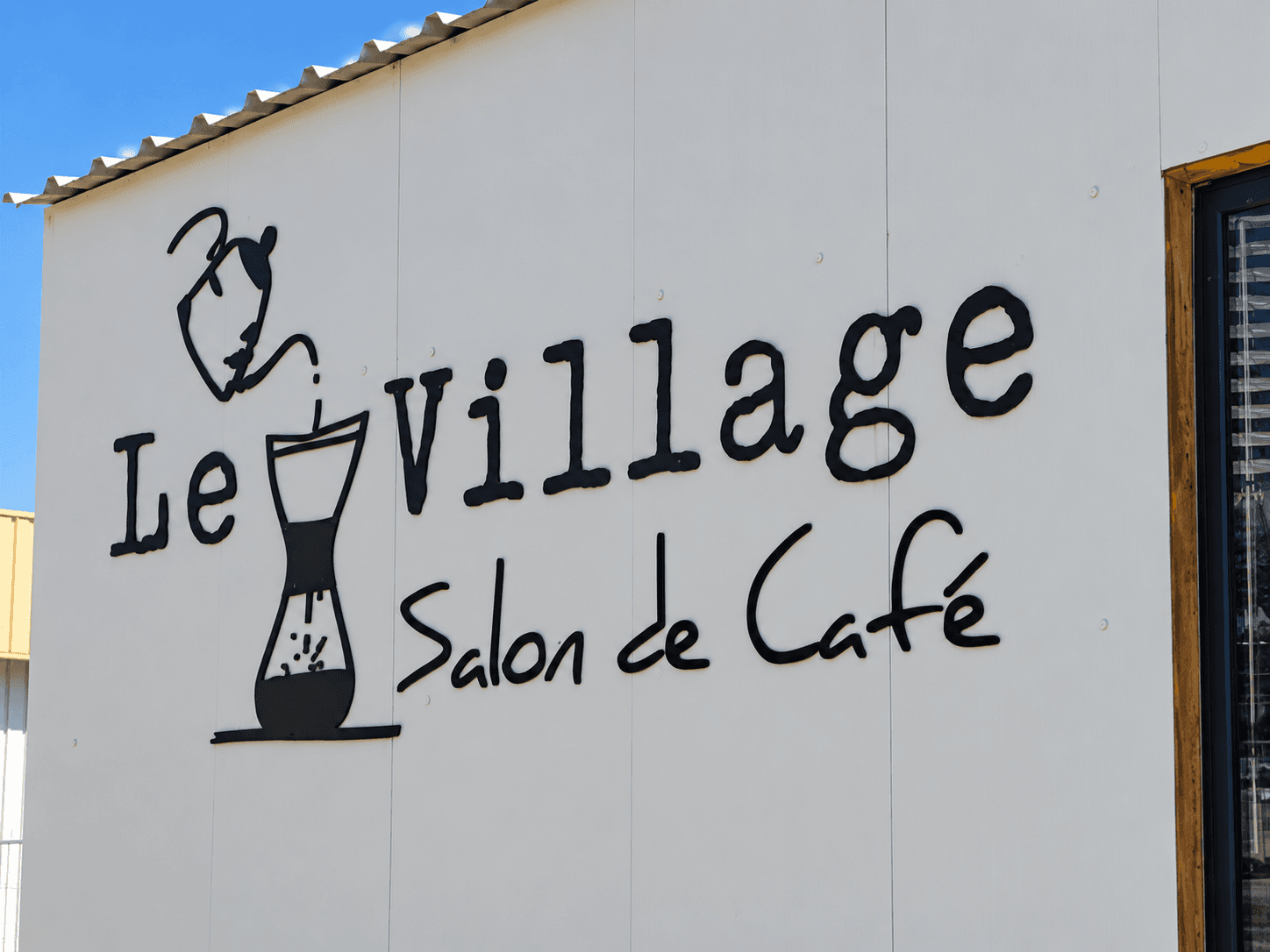 Enseigne extérieure en lettres découpées pour Le Village, salon de café, avec logo et lettrage noir fixés sur la façade du bâtiment.