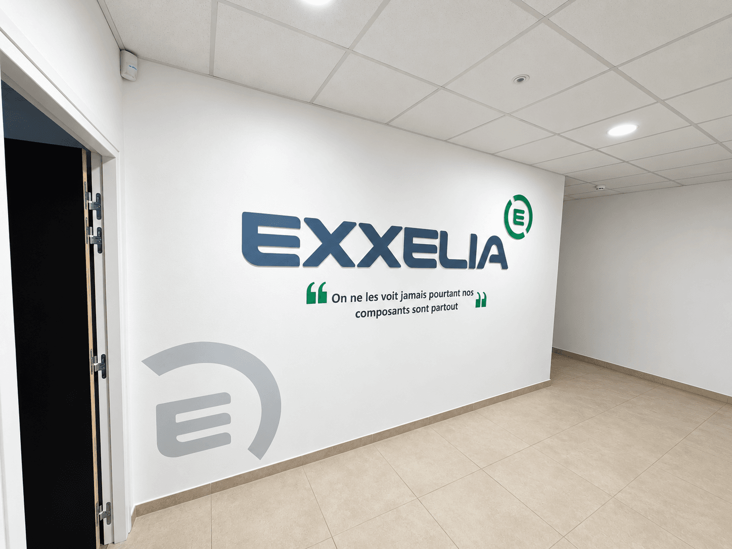 Enseigne intérieure Exxelia en lettres découpées en Forex, accompagnée du logo vert et d’un adhésif mural reprenant la signature de l’entreprise.
