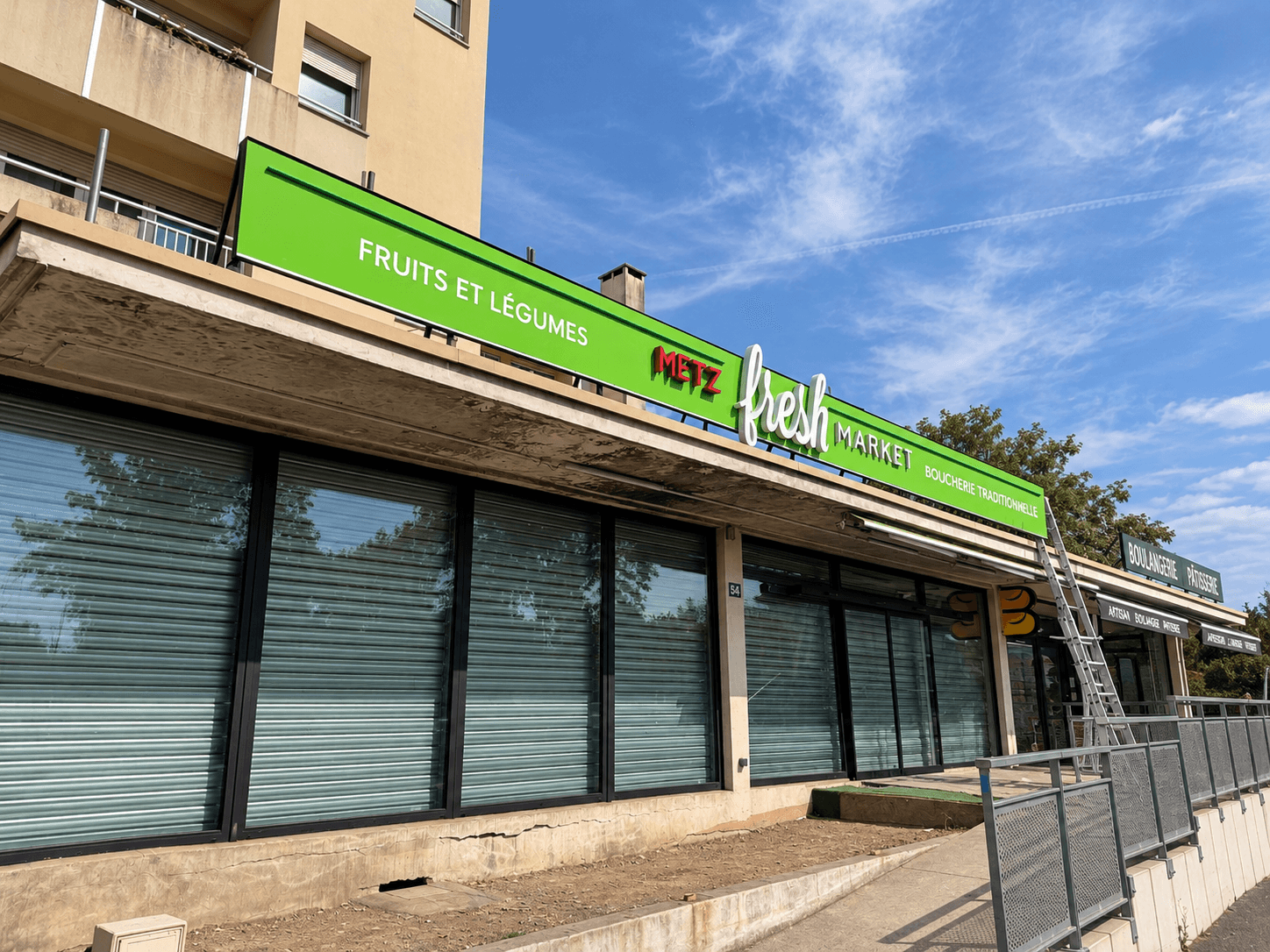 Vue latérale de l’enseigne commerciale Metz Fresh Market après réfection, avec lettrage en relief et remplacement des lettres boîtiers « Best » par « Metz ».
